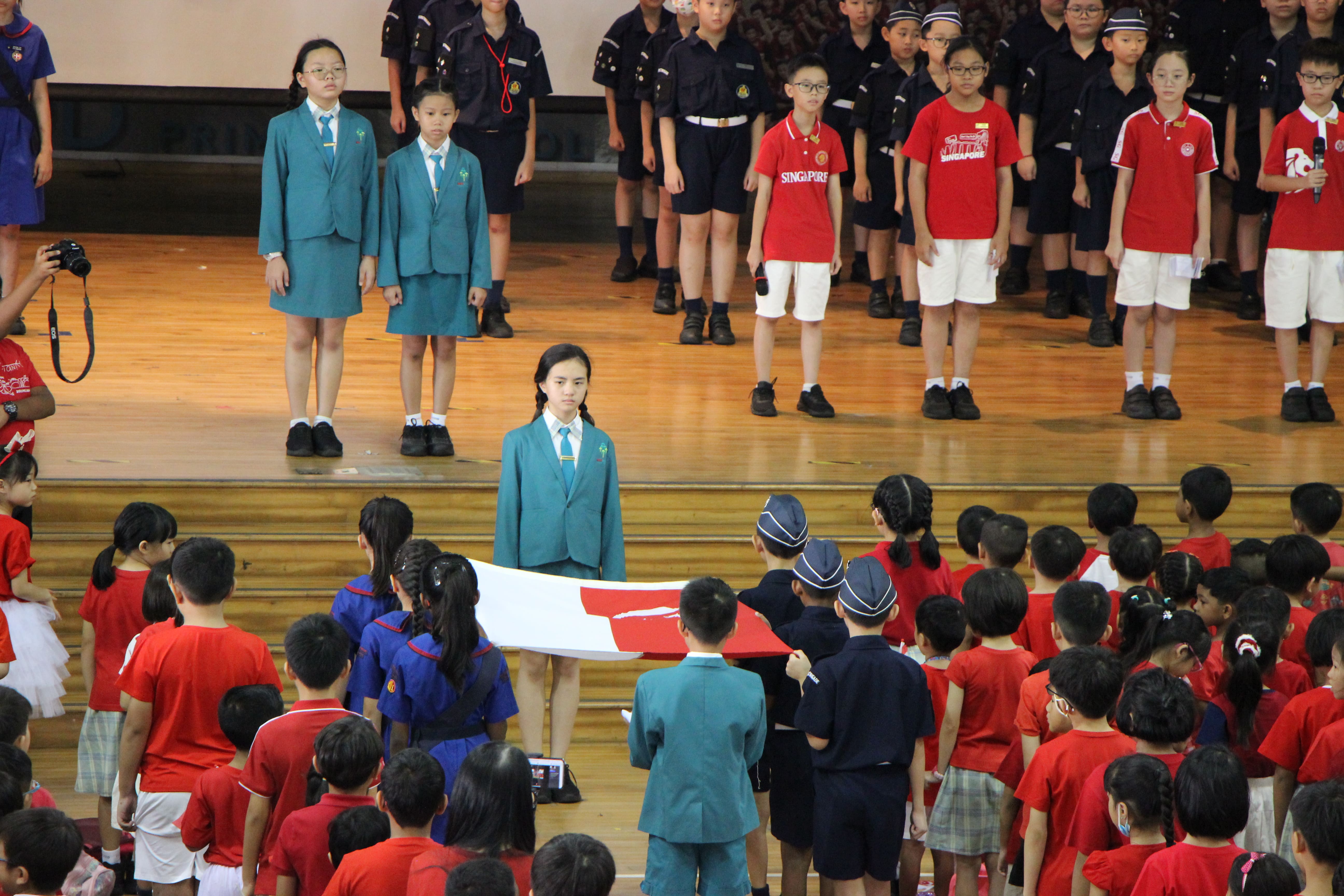 Children in various school uniforms stand at attention during a flag ceremony in a school hall.